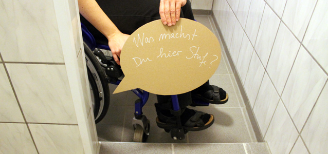 Einjunger Mann im Rollstuhl hält in der Duschkabine ein Schild mit der Aufschrift "Was machst Du hier, Schwelle" in den Händen, um darauf aufmerksam zu machen, dass dies nicht barrierefrei ist.