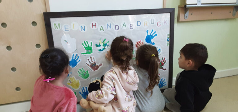 Vier Kinder sitzen vor einem großen Bild mit ihren Handabdrücken und probieren den Bookii-Stift aus.