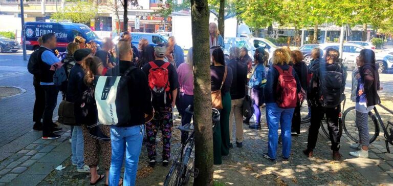 Eine Gruppe vieler Menschen steht an einer Kreuzung neben einem Baum und lauscht den Instruktionen der Ralley-Mitarbeiterin, die auf einem kleinen Podest steht.