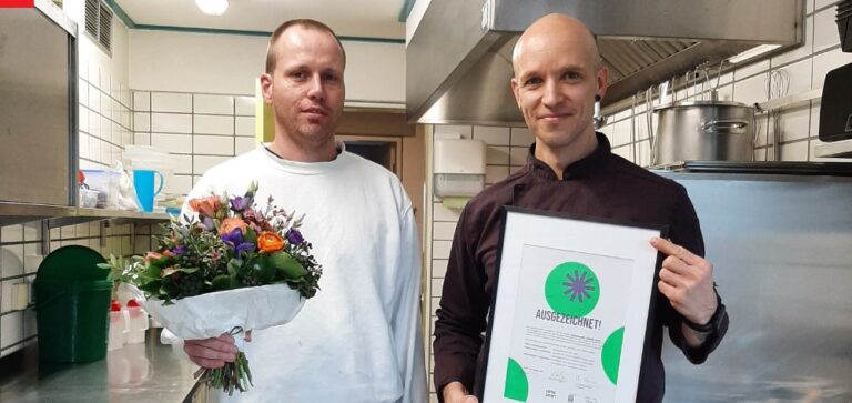 Zwei junge Männer, die zwischen Küchengeräten in einer Kita-Küche stehen, halten eine Urkunde "Kantine Zukunft" und einen Blumenstrauß in der Hand und lächeln in die Kamera.