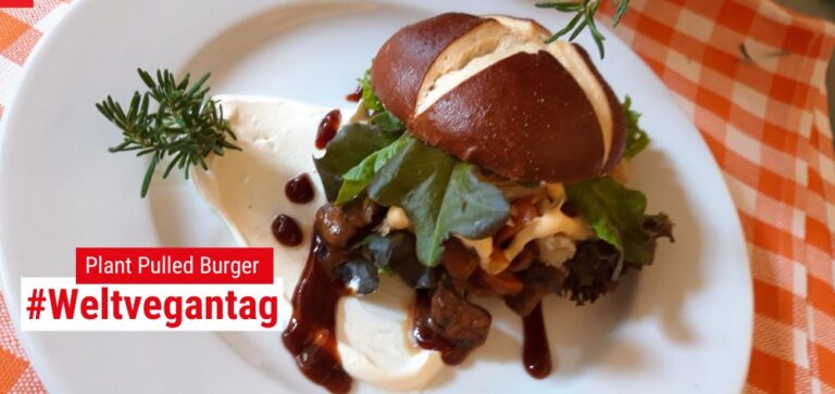 Zwischen einem Laugenbrötchen liegt eine Scheiben aus veganem Hack; darüber Salat und eine Jus tropft heraus. Das Ganze liegt auf einem Teller auf einer rot-weiß-karierten Tischdecke,